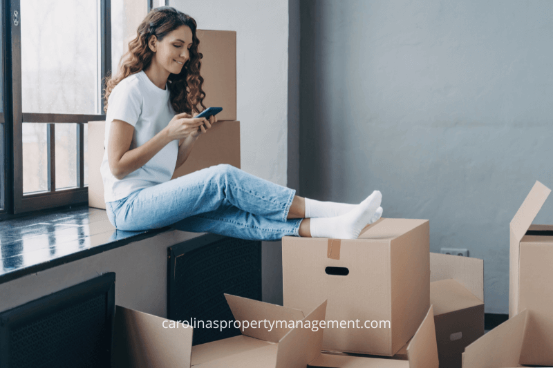 A young woman sitting on moving boxes, smiling while using her phone, surrounded by unpacked boxes in a new home. This image highlights the smooth moving experience provided by Carolina Property Management, a top property management company in Charlotte, NC, helping tenants settle into their ideal rental properties.