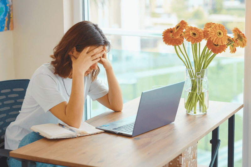 A frustrated landlord sitting at a desk with her hands on her head, staring at a laptop. The workspace includes a notebook and a vase of bright orange flowers by a window. This image illustrates the stress landlords can experience when dealing with complex property management issues, emphasizing how Carolina Property Management LLC offers expert solutions to help landlords manage their properties efficiently and reduce stress.